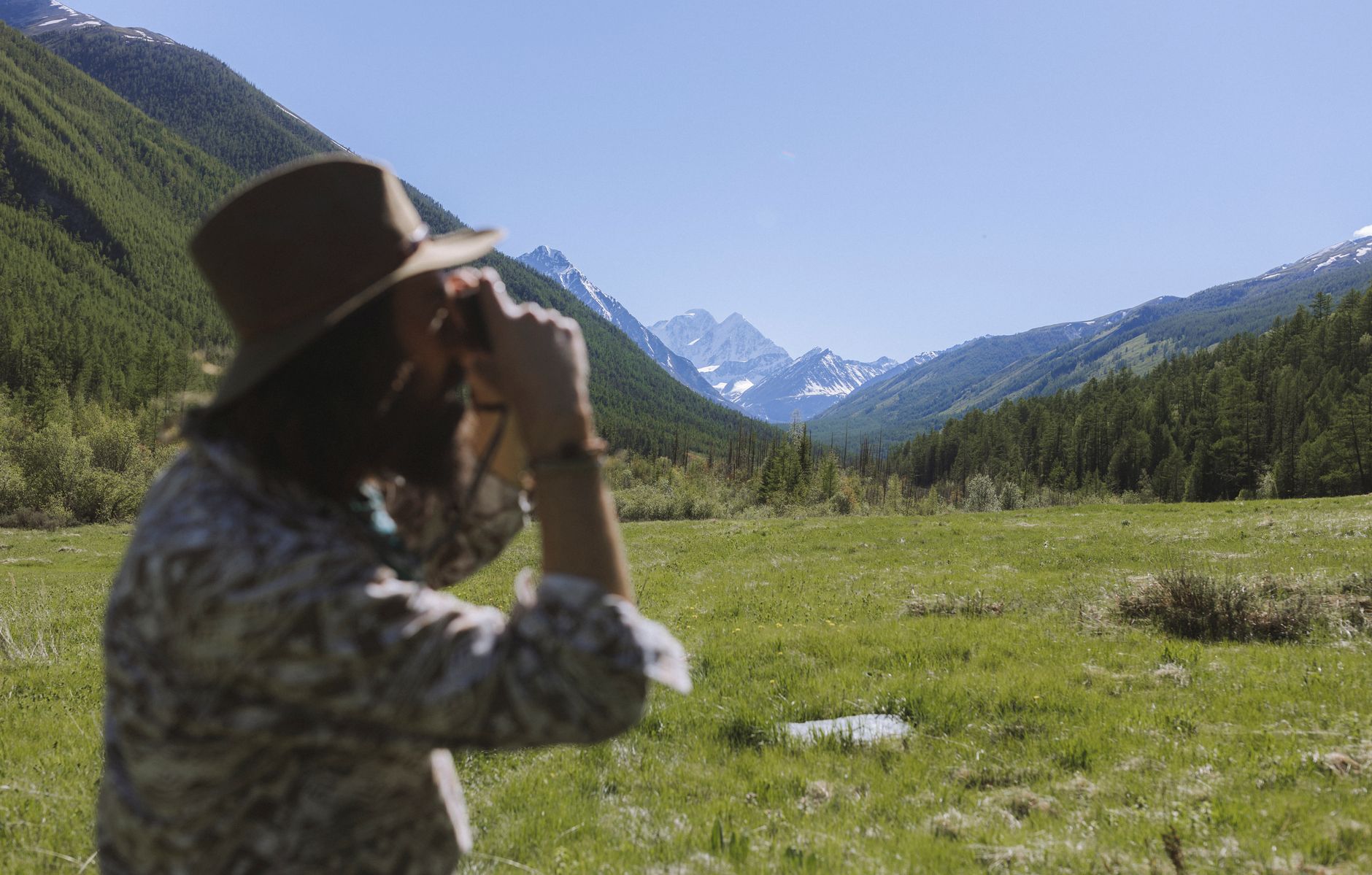 Une personne de dos avec un chapeau regarde à travers des jumelles vers une vallée de montagne.